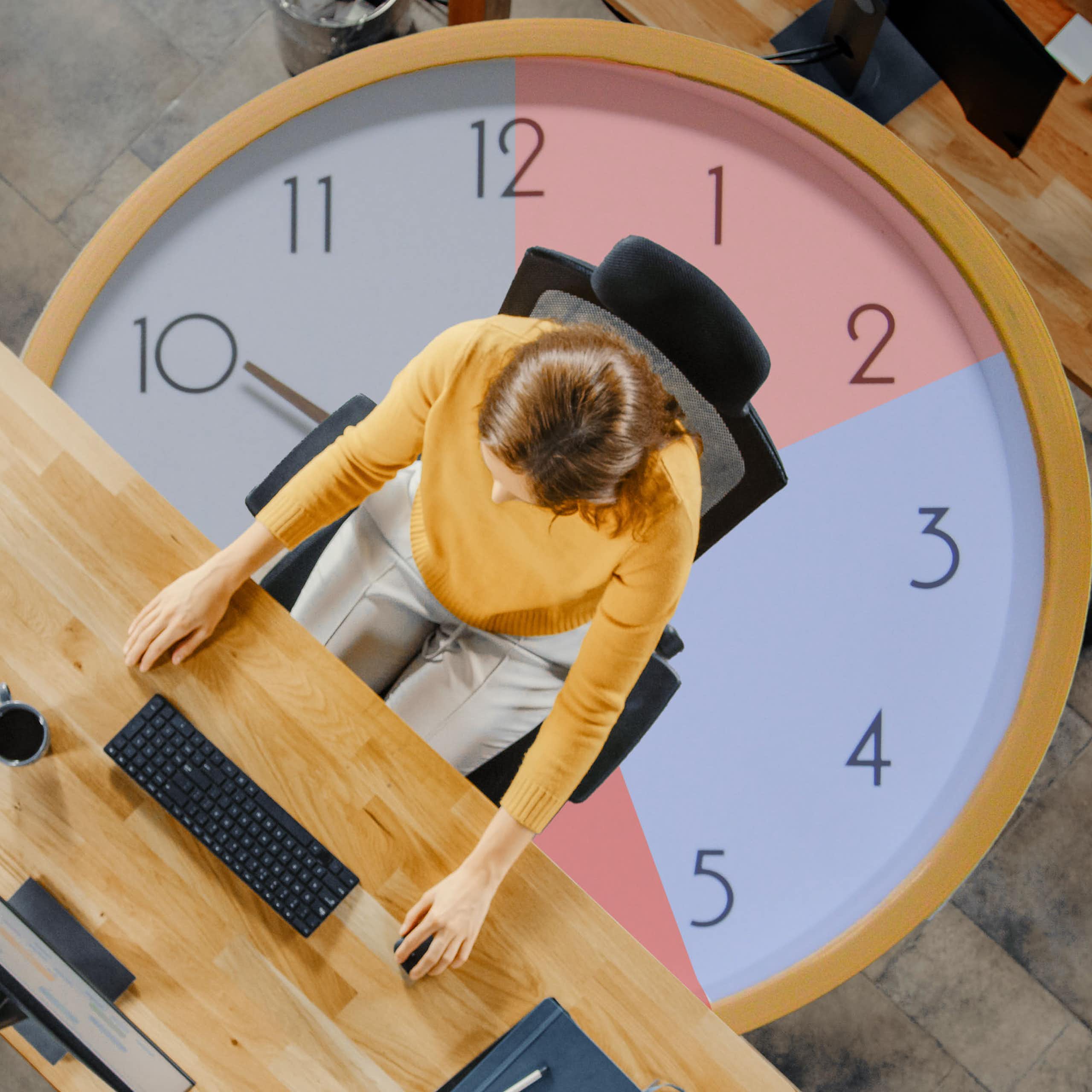 Top view shot of a young woman sitting at her desk working on a computer with a clock in the background.