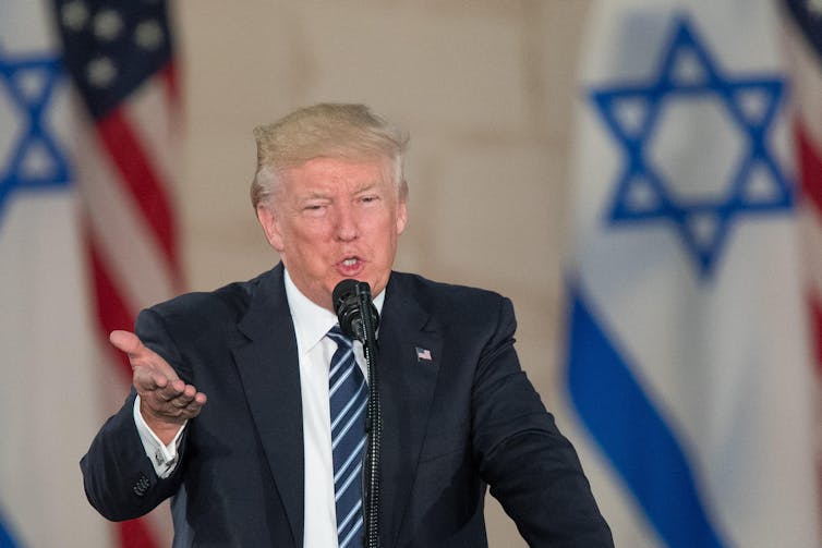 Donald Trump standing in front of the US and Israeli flags.