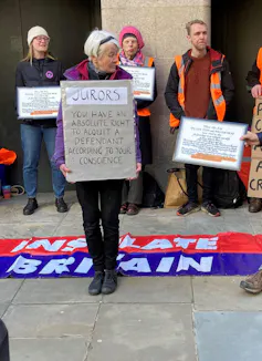 An older woman holding a sign with an Insulate Britain flag on the ground behind her.