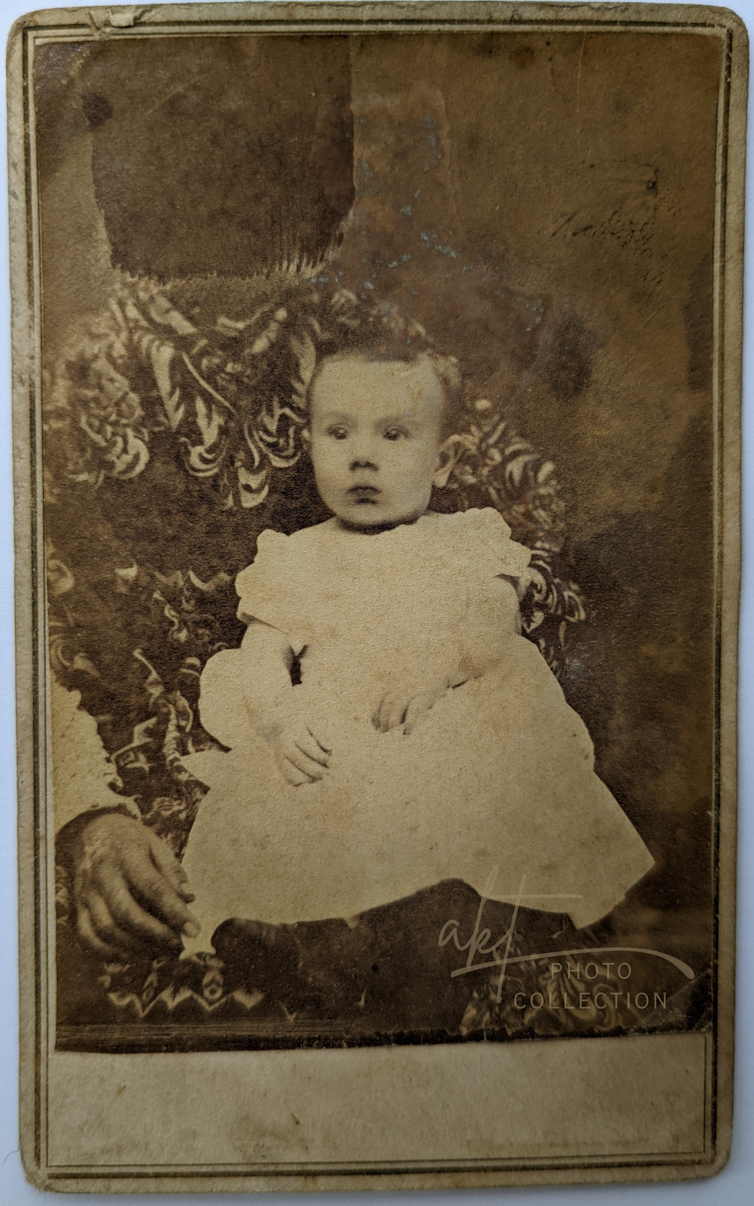 Sepia photo of young child in a dress, held in the lap of an adult whose face is covered by a black box