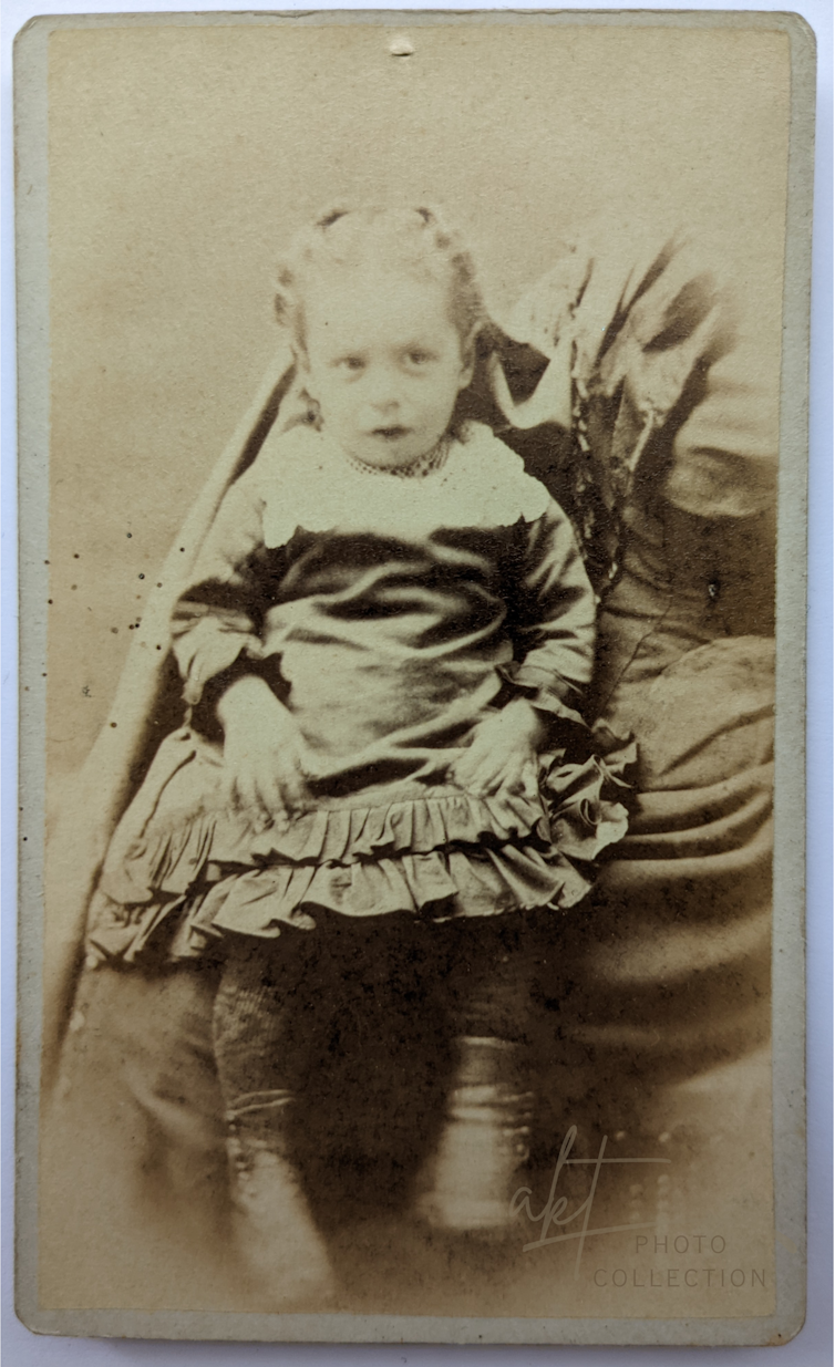 Sepia photograph of toddler in dress sitting in lap of adult with cut off head and legs