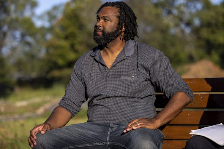 A man with black hair and a beard sitting on a bench outside.