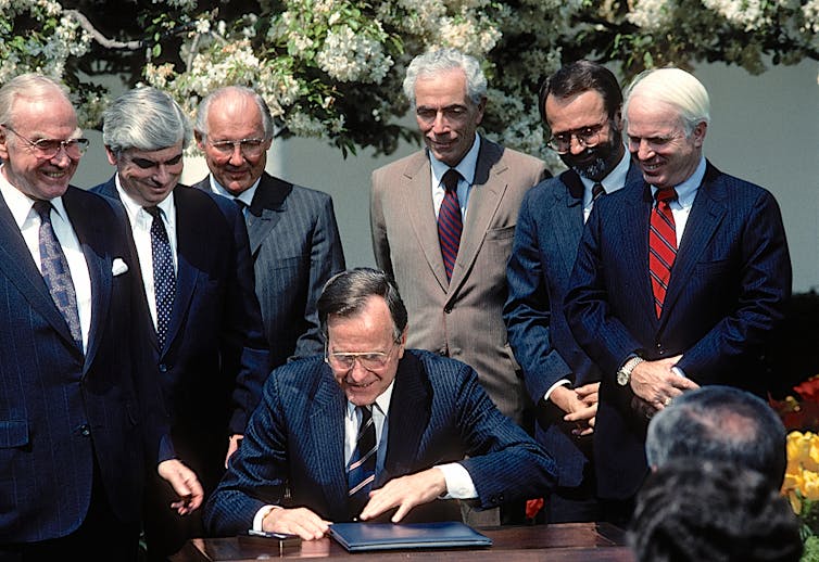 Un grupo de hombres trajeados rodean a un hombre trajeado sentado frente a un escritorio.  Todos ellos están sonriendo.
