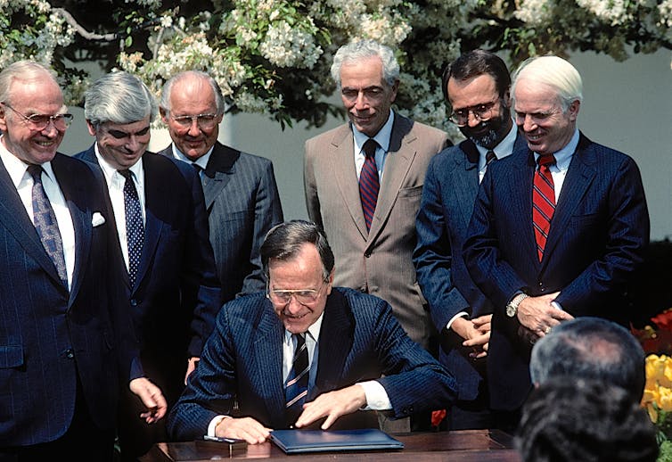 A group of men in suits stand around a man in a suit sitting at a desk. All of them are smiling.