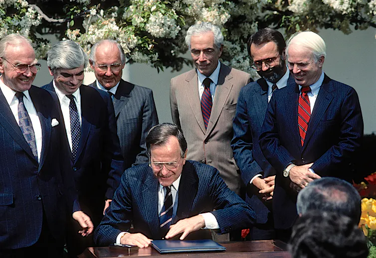 A group of men in suits stand around a man in a suit sitting at a desk. All of them are smiling.