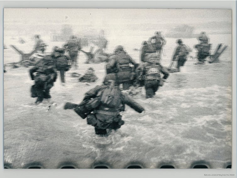 A slightly blurry black and white image behind a group of soldiers, showing them stepping into waist-deep seawater.