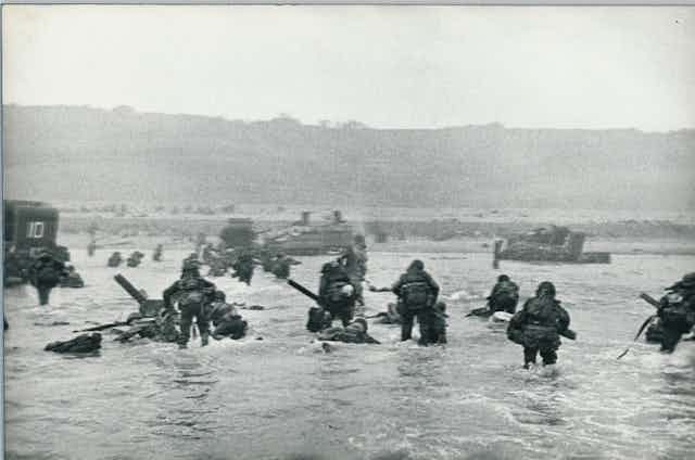 A black-and-white image shows soldiers wading through thigh-deep water toward a beach backed by cliffs.