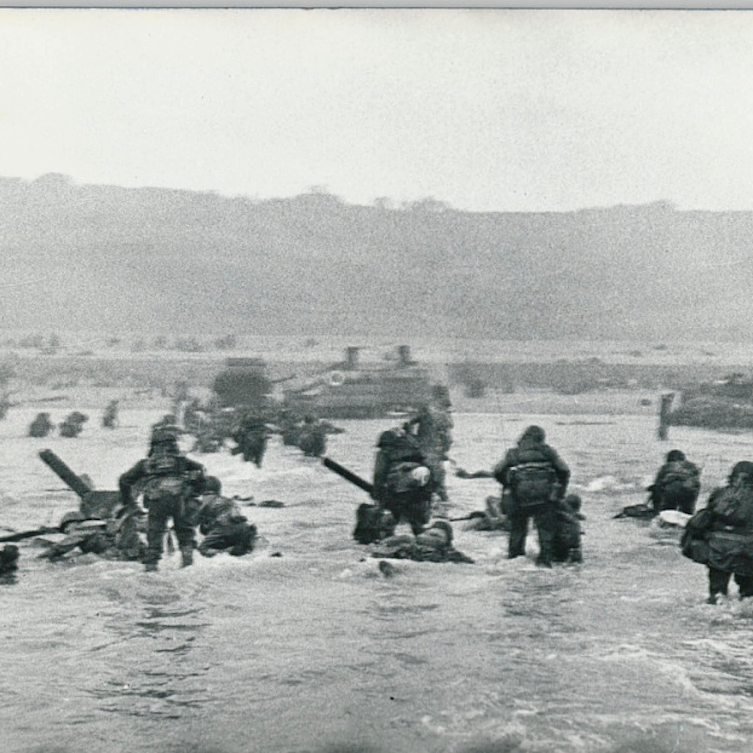 A black-and-white image shows soldiers wading through thigh-deep water toward a beach backed by cliffs.