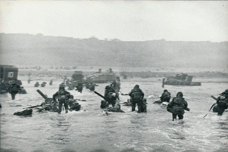 A black-and-white image shows soldiers wading through thigh-deep water toward a beach backed by cliffs.