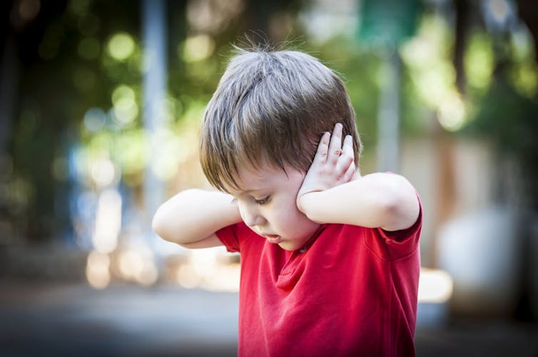 Small boy standing outside, eyes down, hands over ears