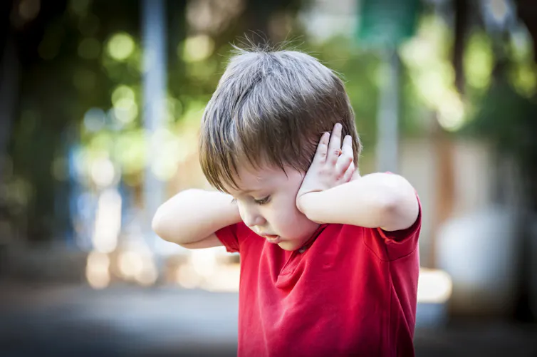 Small boy standing outside, eyes down, hands over ears