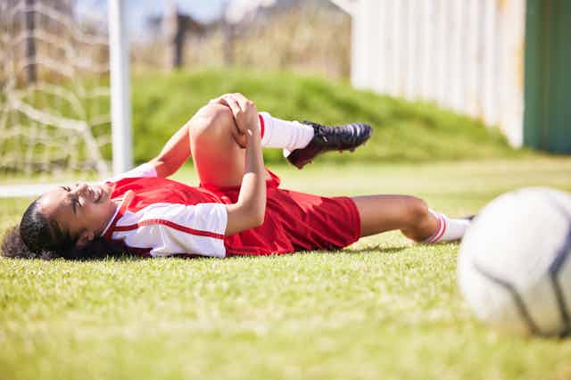 A young woman lying on a soccer pitch clutching her knee in pain.