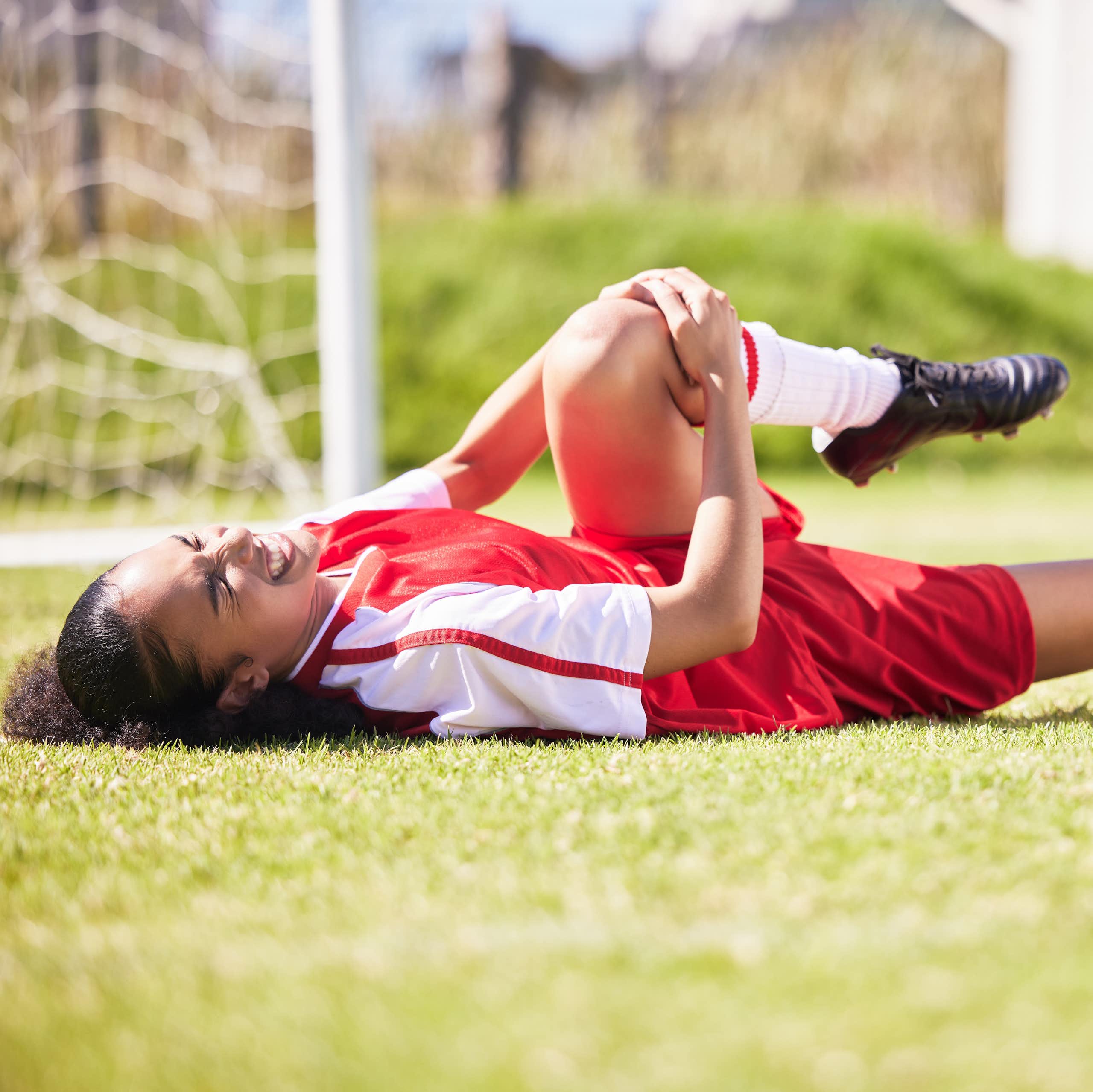 A young woman lying on a soccer pitch clutching her knee in pain.
