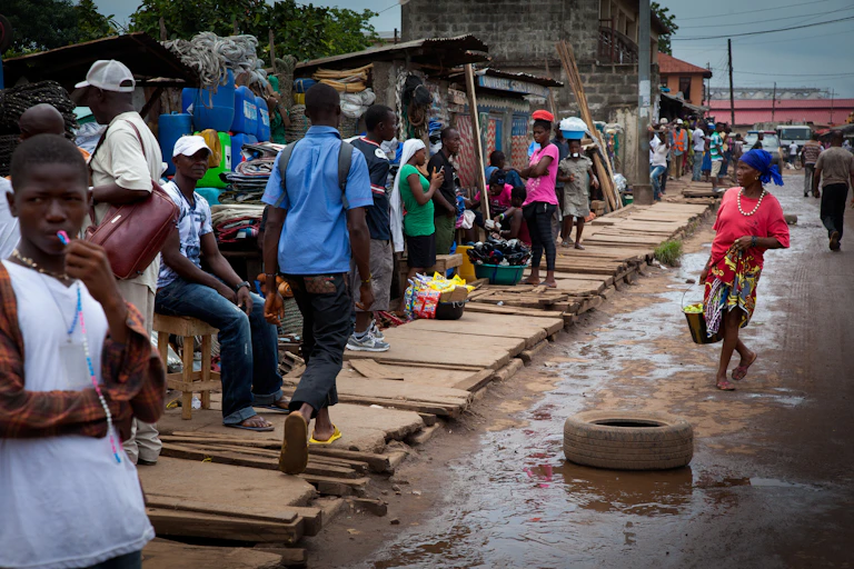 People in a muddy street, some apparently selling goods at makeshift stalls, others walking past