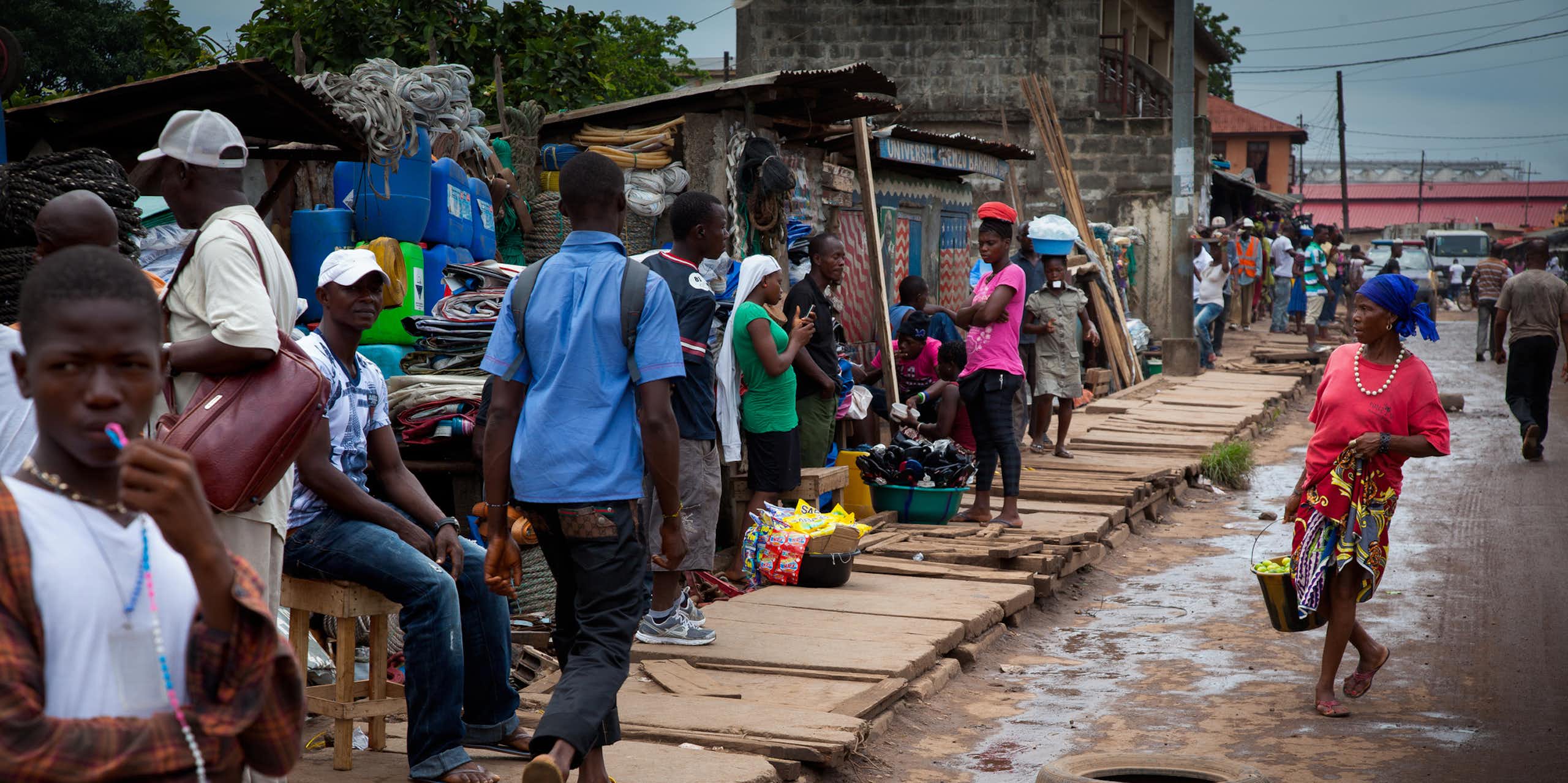 People in a muddy street, some apparently selling goods at makeshift stalls, others walking past