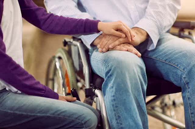 Close up of a man and woman sitting side by side, he is in a wheelchair and she is resting her hand on his, their faces are not visible in the photo
