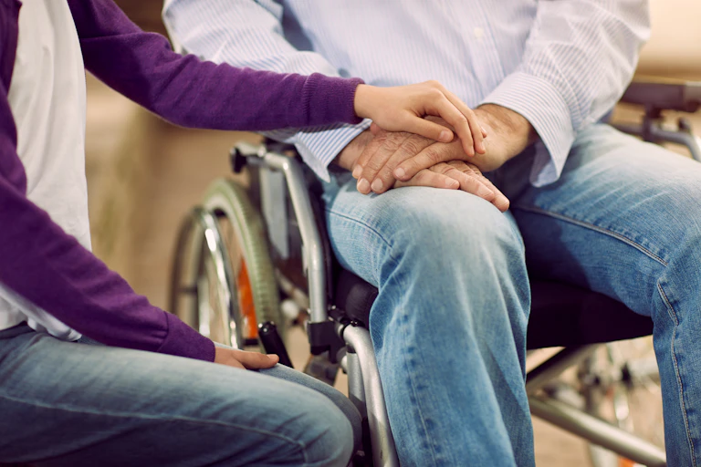 Close up of a man and woman sitting side by side, he is in a wheelchair and she is resting her hand on his, their faces are not visible in the photo