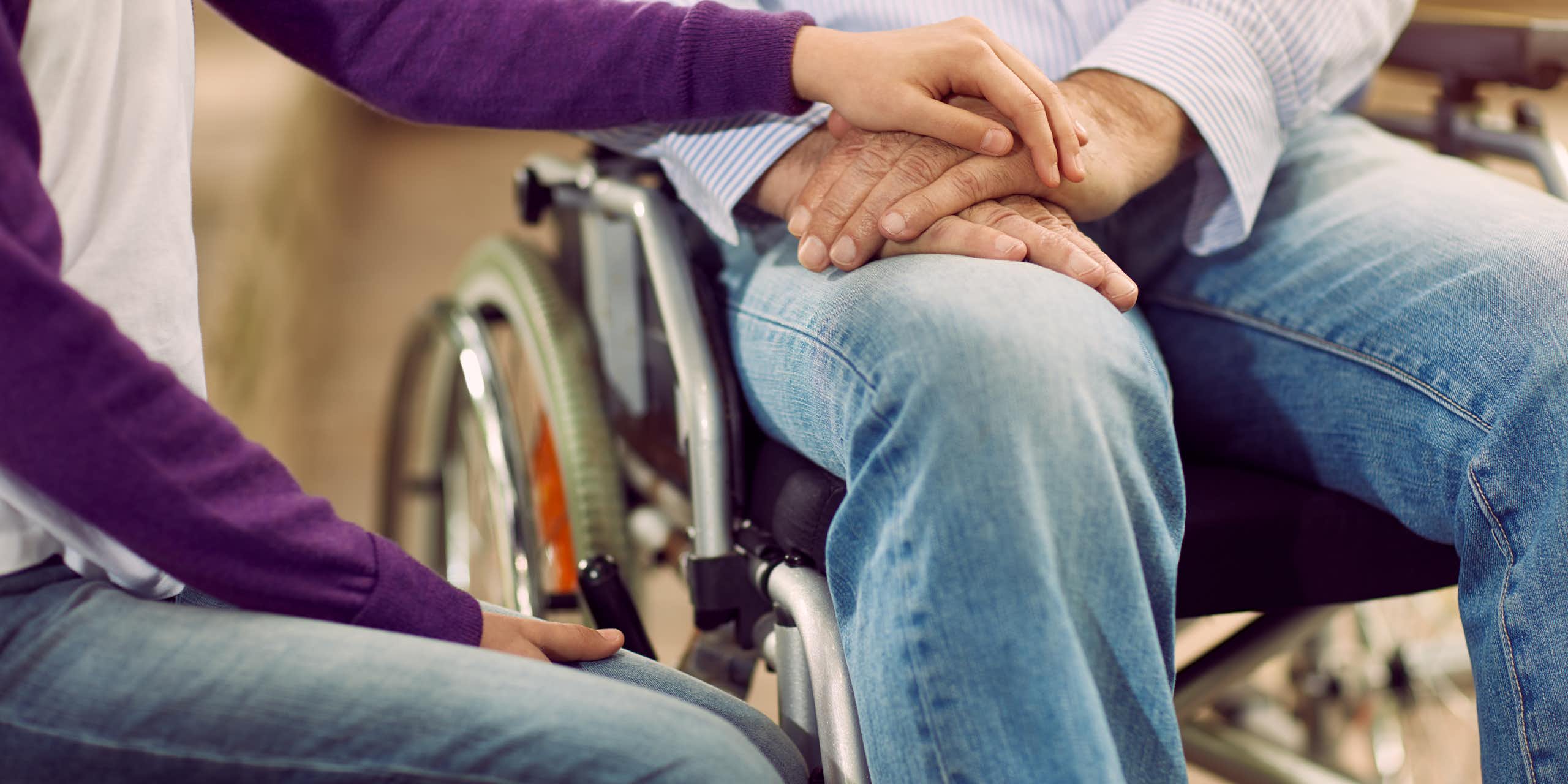 Close up of a man and woman sitting side by side, he is in a wheelchair and she is resting her hand on his, their faces are not visible in the photo