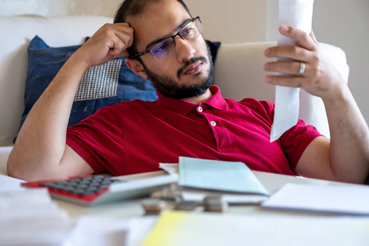 A young man sitting slumped against a couch, frustrated and resting his head on his hand while he looks at a receipt