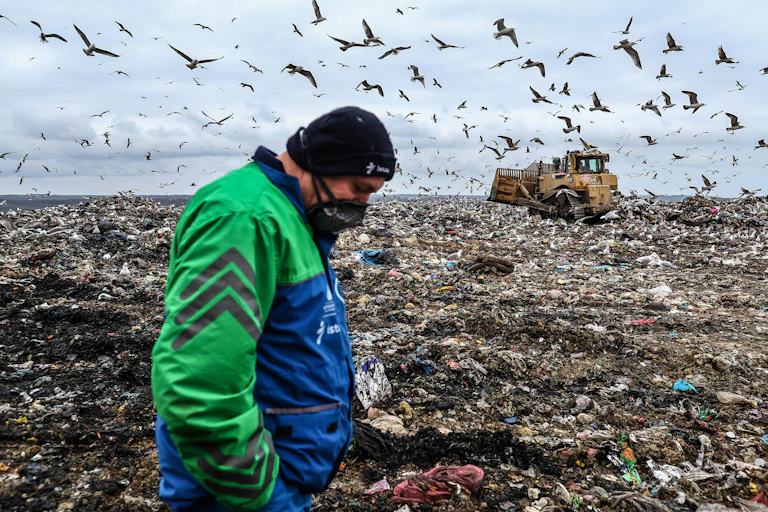 A worker in protective mask walks across a waste dump with birds and a bulldozer in the background.