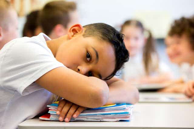 Elementary school-age boy rests head on crossed arms on desk with other kids in background