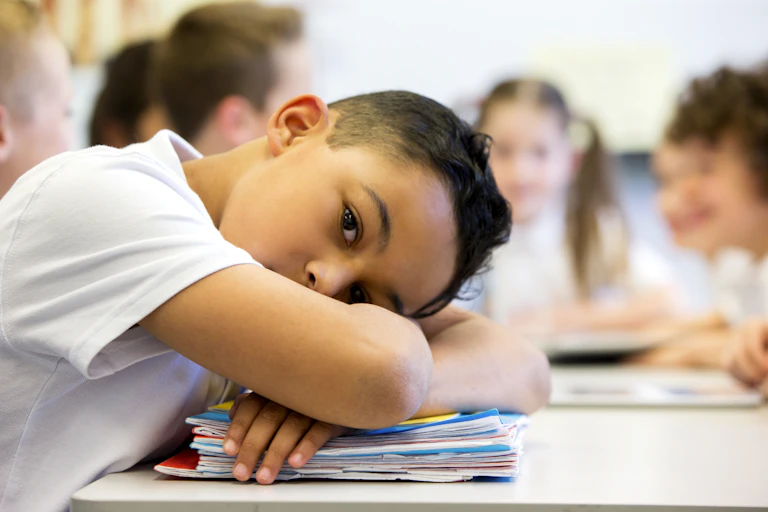 Elementary school-age boy rests head on crossed arms on desk with other kids in background