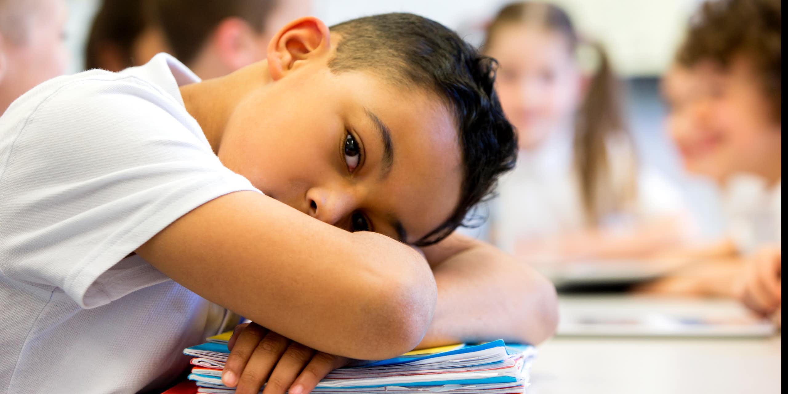 Elementary school-age boy rests head on crossed arms on desk with other kids in background