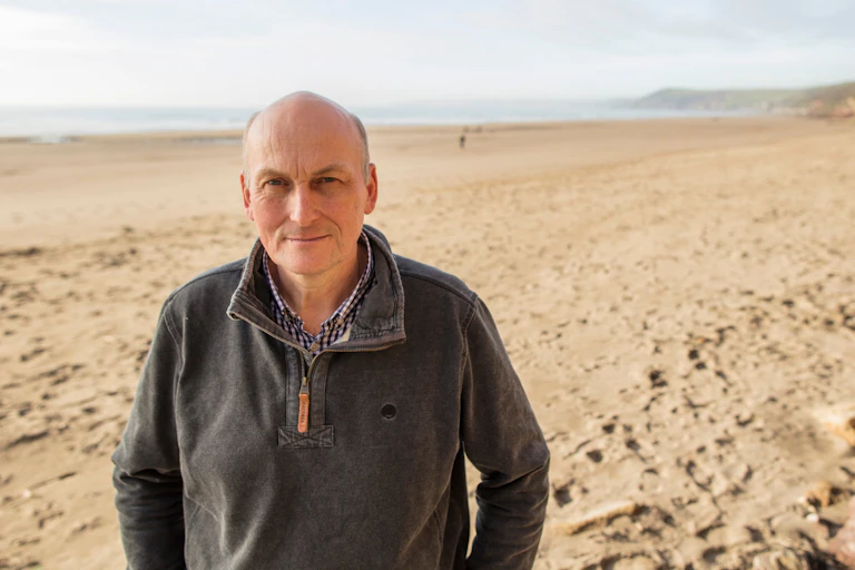 Man in grey jumper facing camera, wide empty sandy beach and sea in background