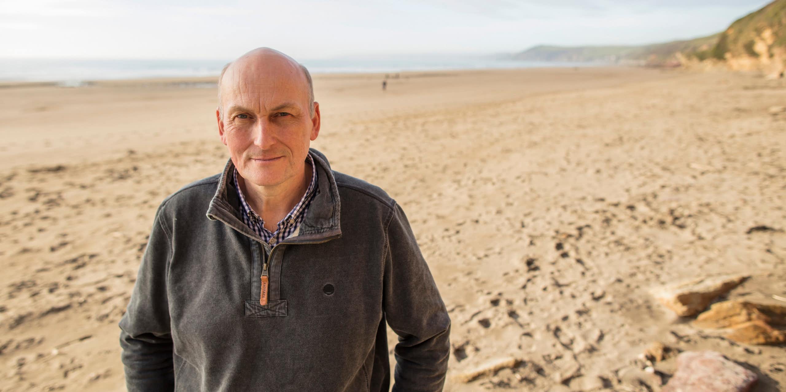 Man in grey jumper facing camera, wide empty sandy beach and sea in background
