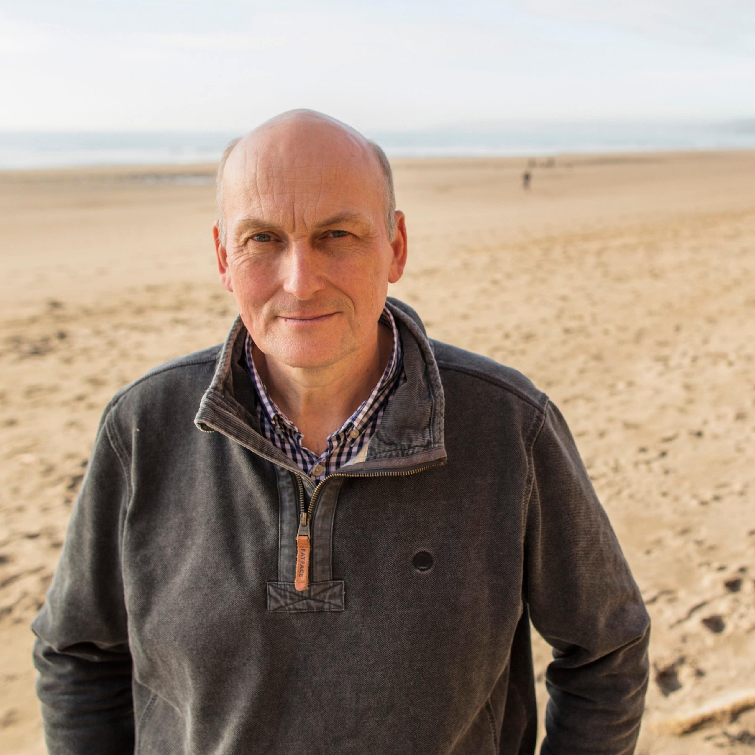 Man in grey jumper facing camera, wide empty sandy beach and sea in background