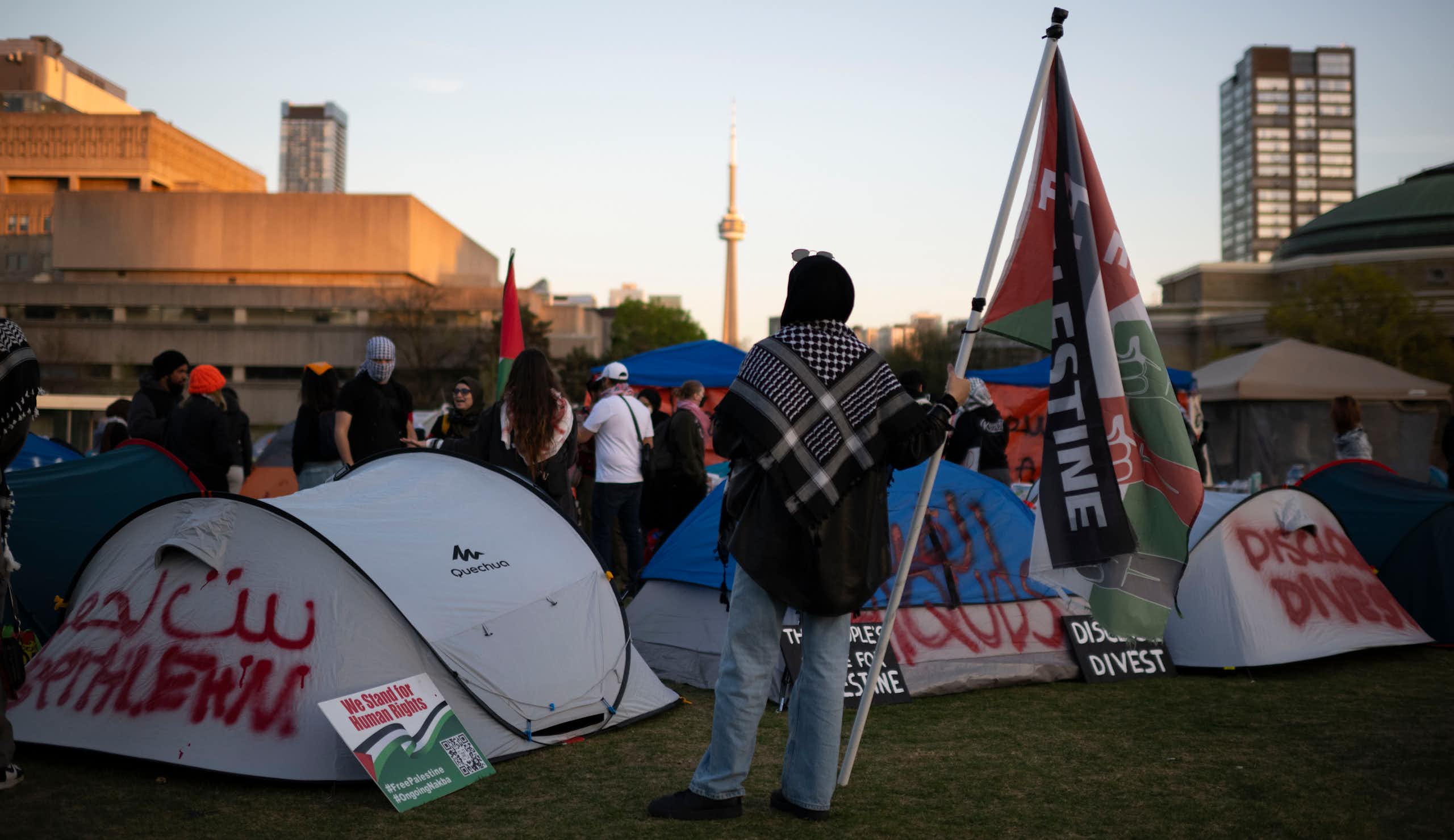 A person seen holding a flag with the end of the word 'Palestine' visible against tents and a Toronto skyline including the CN tower.