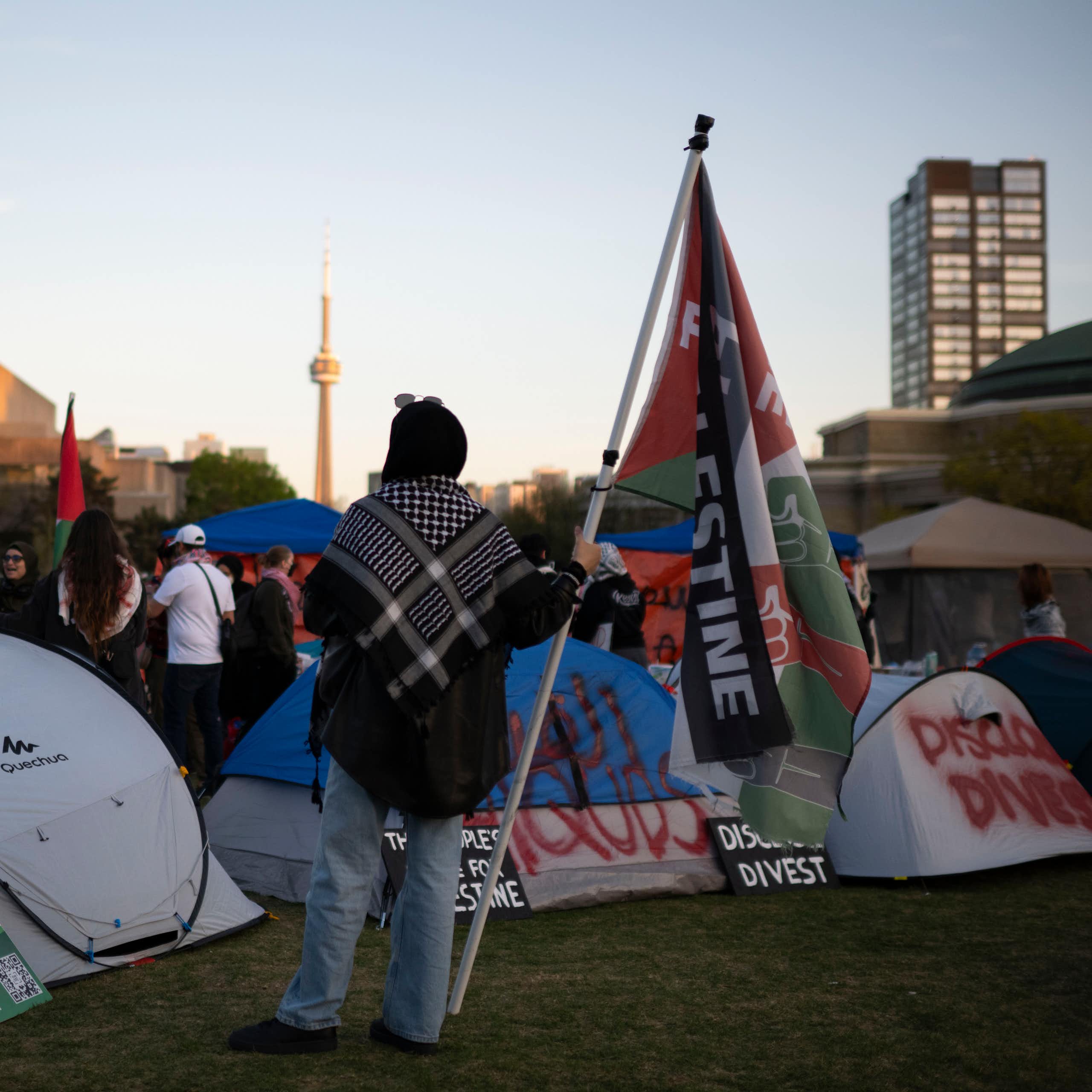 A person seen holding a flag with the end of the word 'Palestine' visible against tents and a Toronto skyline including the CN tower.