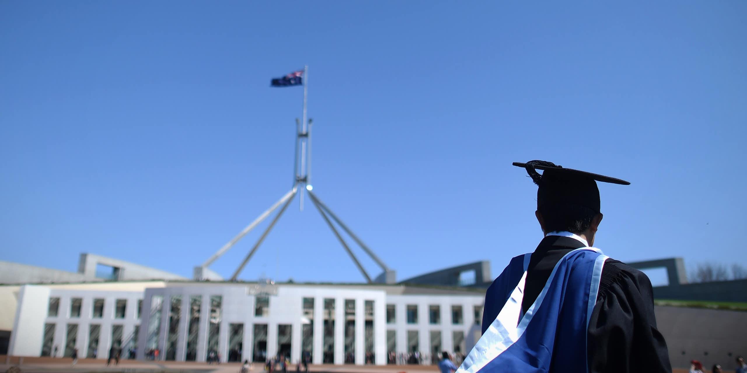 A graduate in cap and gown stands in front of federal parliament. 