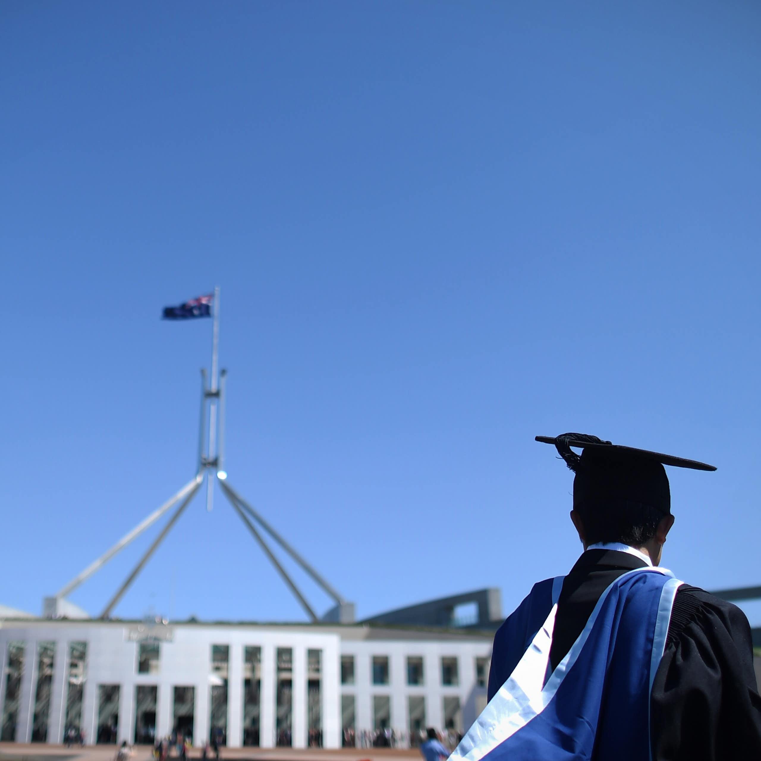 A graduate in cap and gown stands in front of federal parliament. 