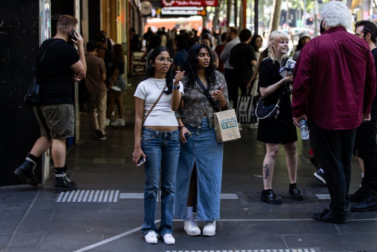 People walk along Swanston Street in Melbourne. Two young women stop at a crossing, carrying a shopping bag.