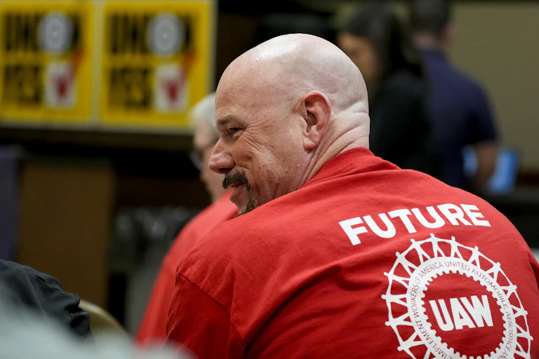 Man smiles while wearing a red t-shirt that ays 'Future UAW' with blurry signs in the background that say 'Union yes' with checkmarks.