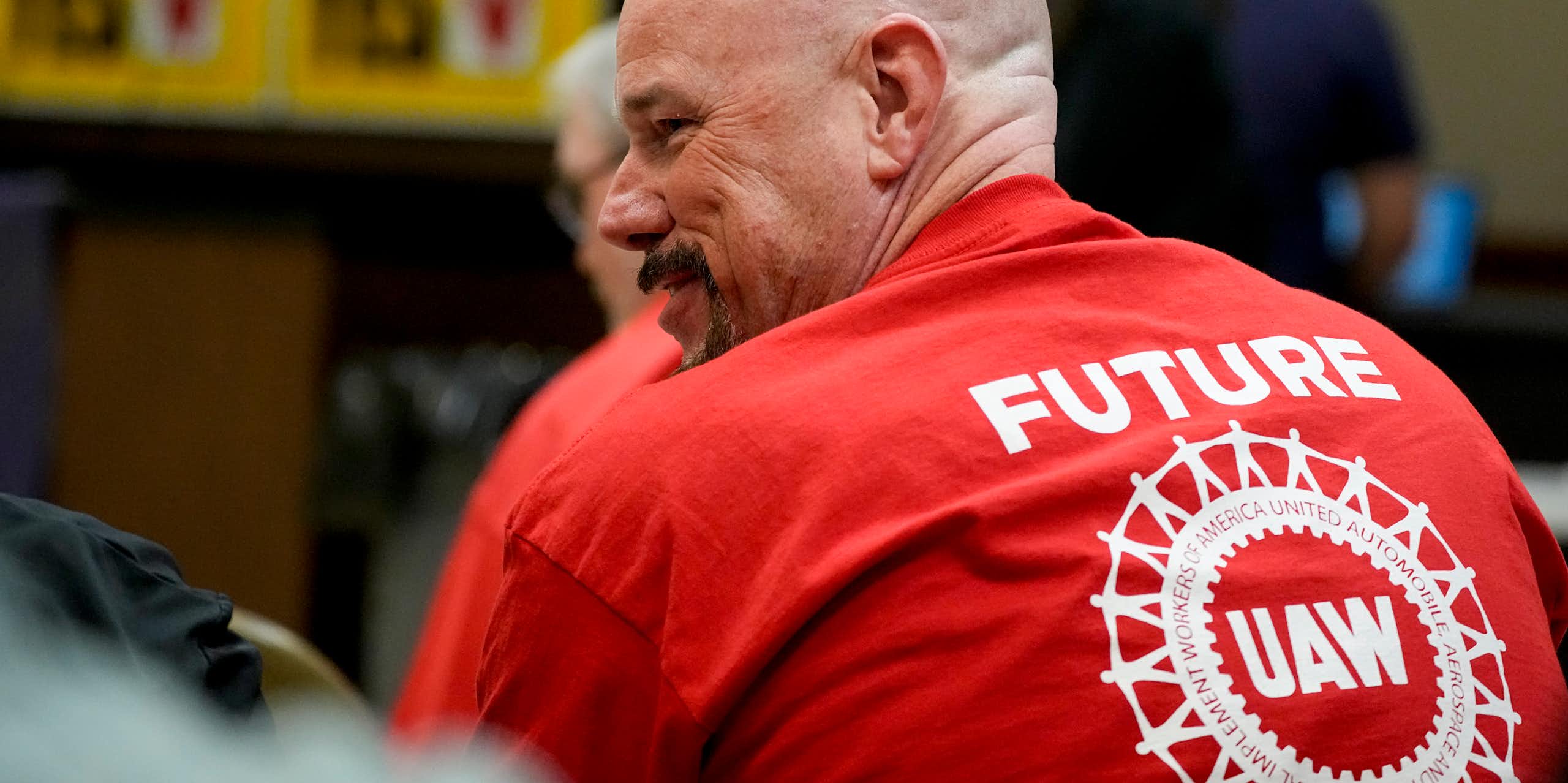 Man smiles while wearing a red t-shirt that ays 'Future UAW' with blurry signs in the background that say 'Union yes' with checkmarks.
