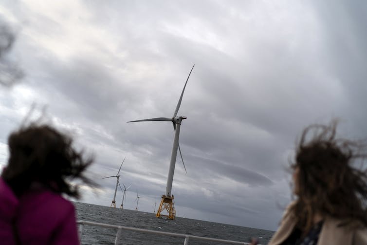 Two people with their hair blowing in the wind look out at a small wind farm with very large wind turbines.