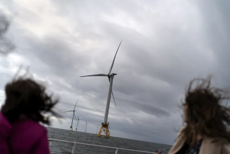 Two people with their hair blowing in the wind look out at a small wind farm with very large wind turbines.