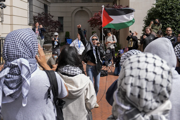 Mujer con gafas de sol ondea una bandera palestina roja, verde, blanca y negra rodeada de gente con keffiyah.
