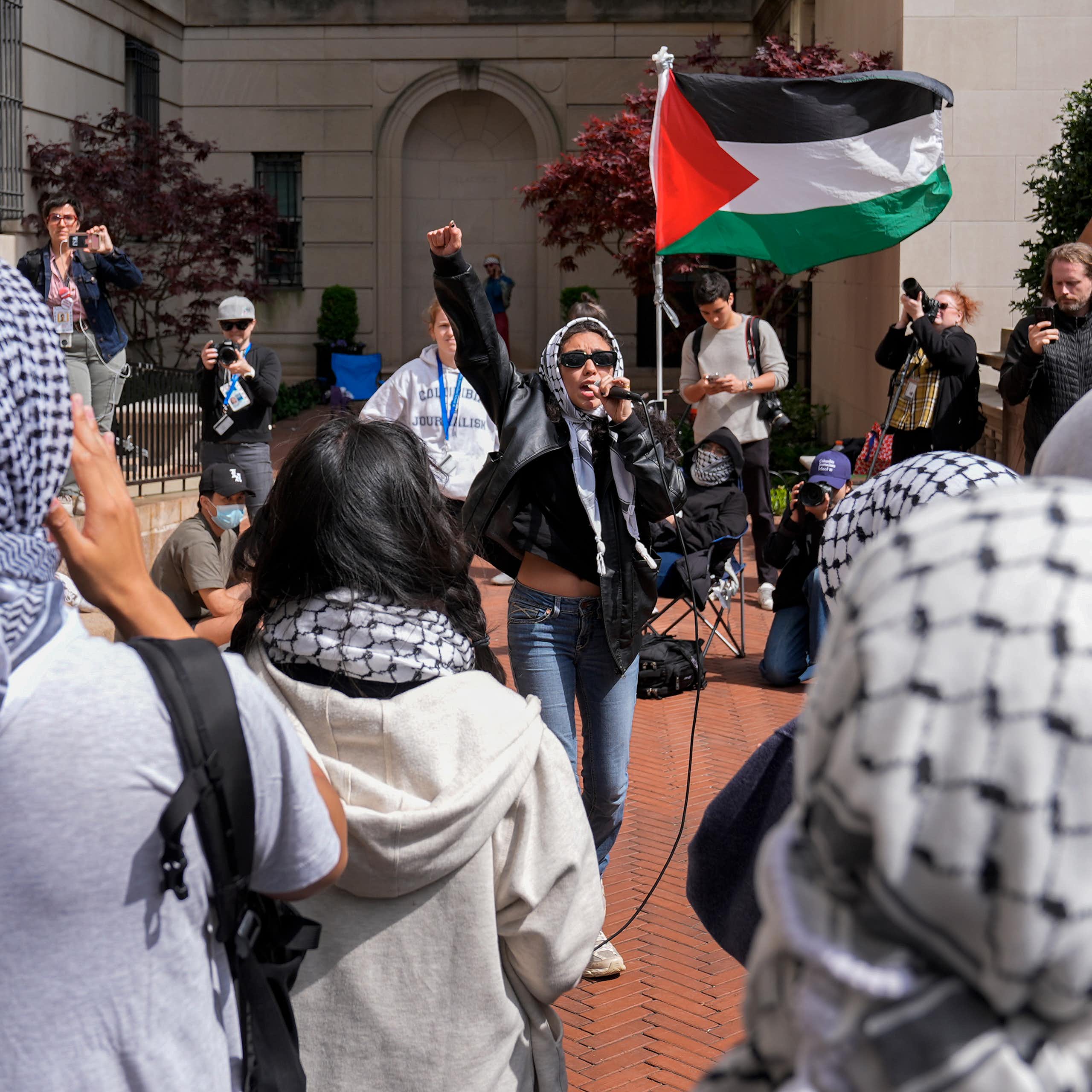 Mujer con gafas de sol ondea una bandera palestina roja, verde, blanca y negra rodeada de gente con keffiyah.