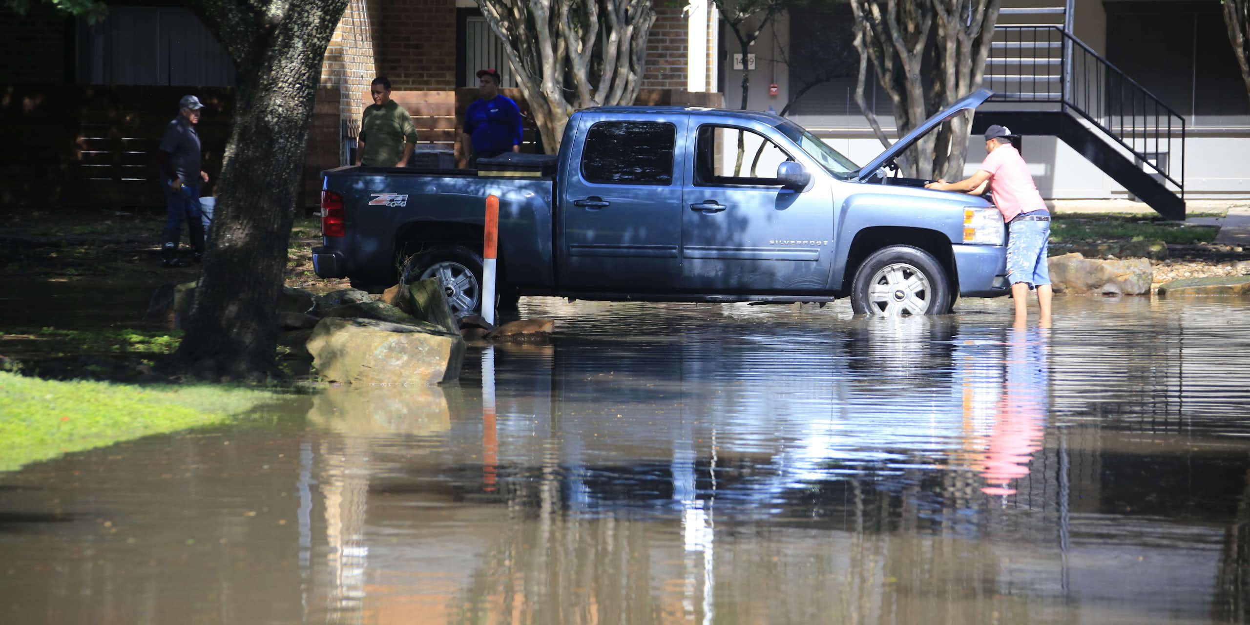 A man works on the engine of a truck while standing in floodwater over his ankles outside a home.