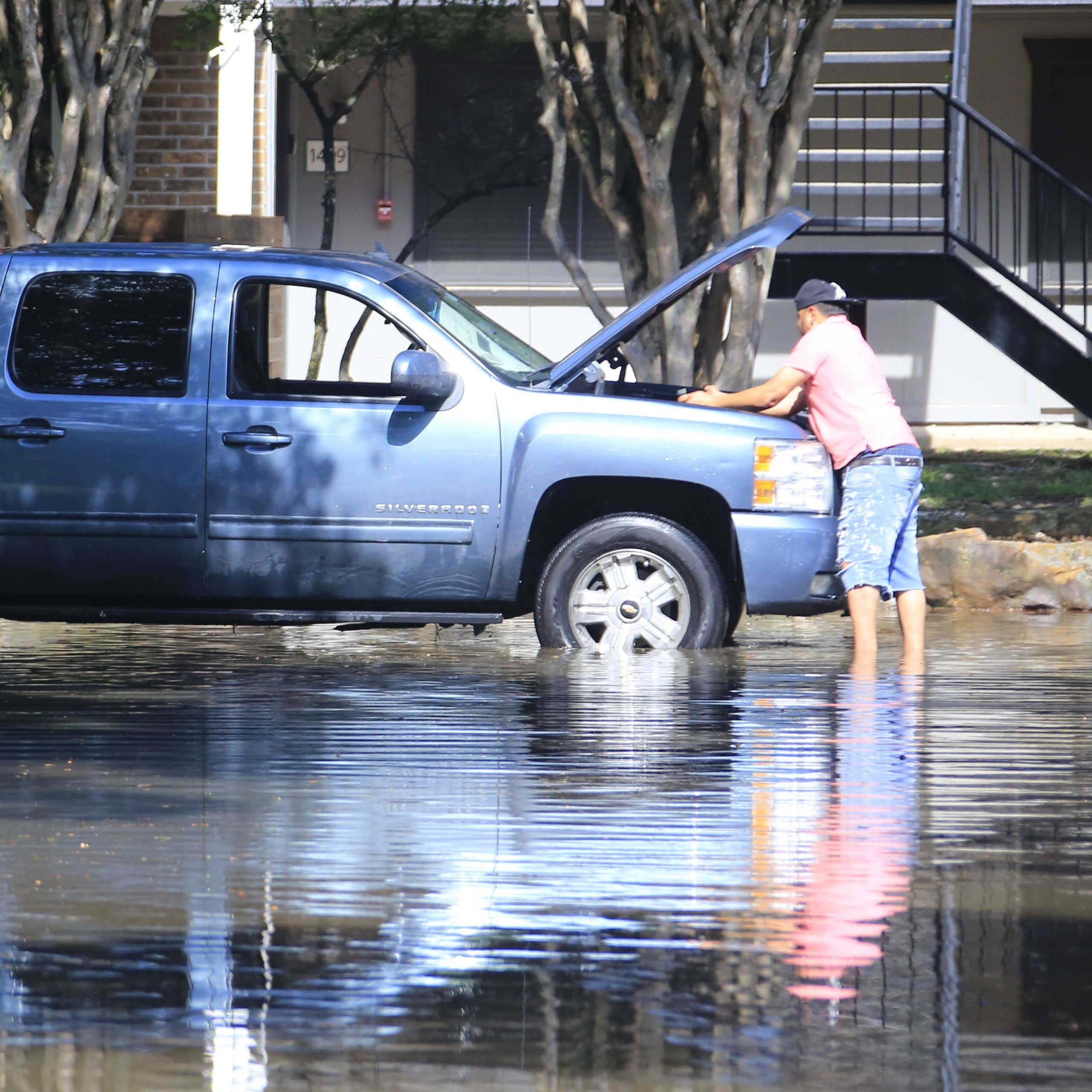 A man works on the engine of a truck while standing in floodwater over his ankles outside a home.