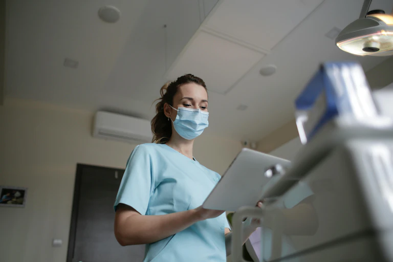 A woman in a surgical mask and scrubs consults a screen in a consulting room.