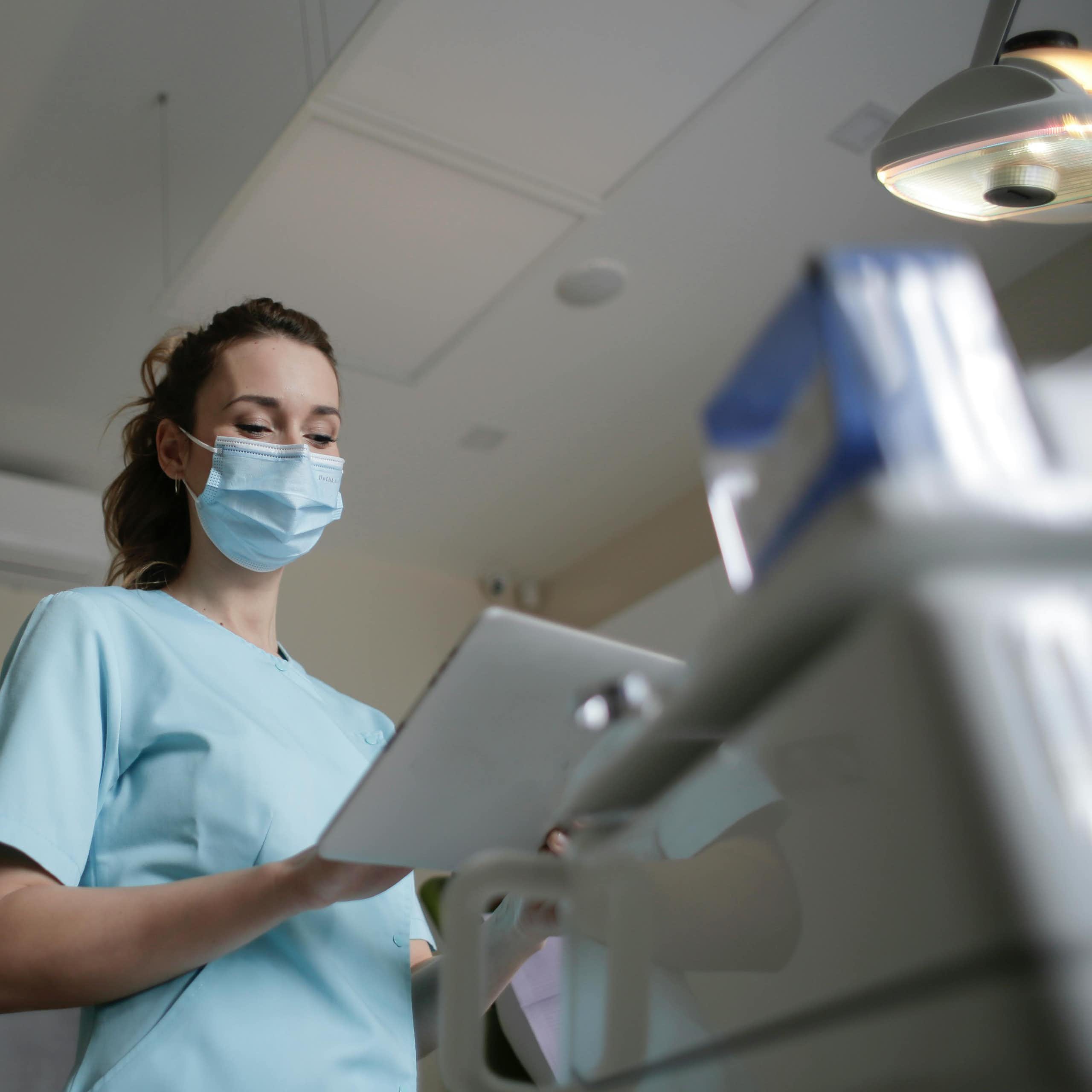 A woman in a surgical mask and scrubs consults a screen in a consulting room. 