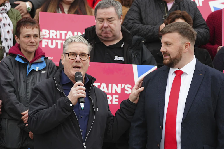 A middle-aged man with glasses speaks into a microphone next to a younger man in a suit and red tie
