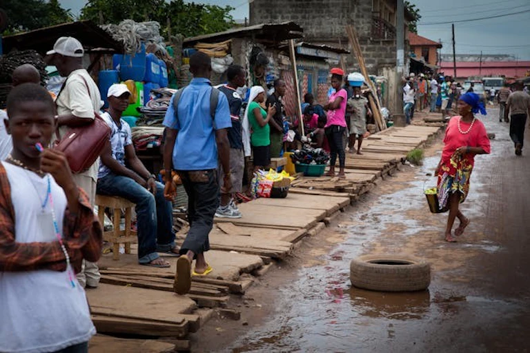 Personnes dans une rue boueuse, certaines vendant apparemment des marchandises sur des étals de fortune, d'autres passant devant.