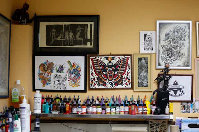 Photo d’un studio de tatouage avec des œuvres d'art encadrées au mur et une table sur laquelle sont alignées des rangées de petites bouteilles d'encre colorée. 
