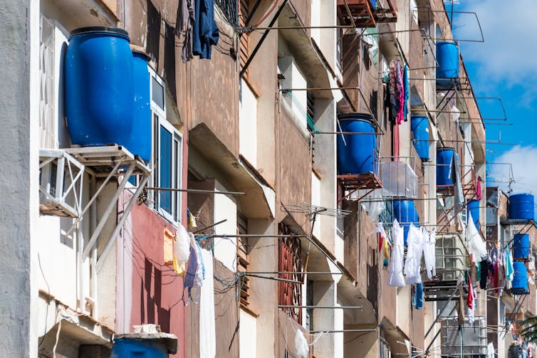 Un edificio de apartamentos con grandes barriles azules de agua situados fuera de cada unidad en plataformas similares a donde se puede ver un aire acondicionado de ventana.