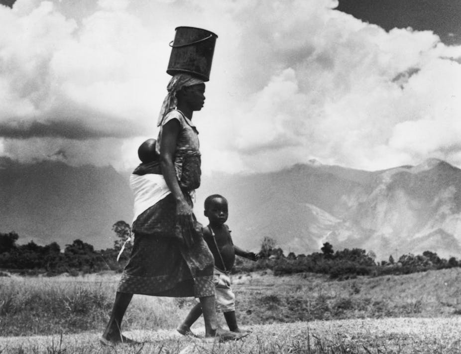 woman walks with water bucket on her head, baby strapped to her back and a young child at her side